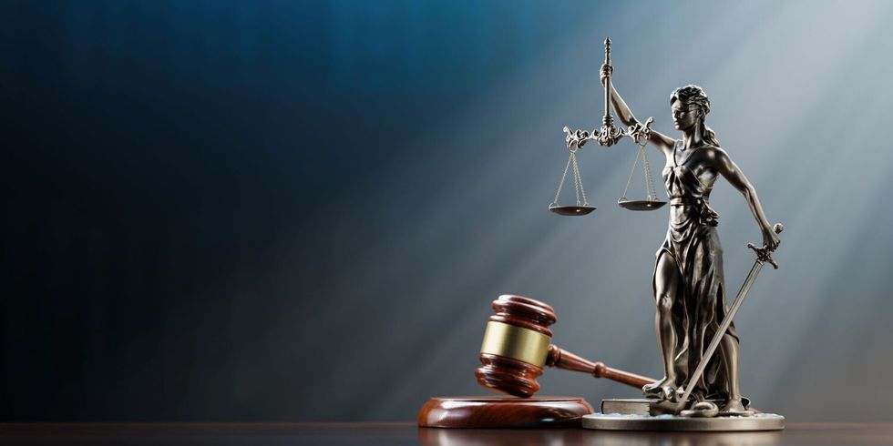 Eine weibliche Figur (genannt Justitia) in langem Gewand mit verbundenen Augen. In der einen Hand hält sie eine Waage, in der anderen ein gesenktes Schwert. Die Darstellung steht für Unparteilichkeit, Abwägung und Durchsetzung des Rechts. Daneben liegt ein brauner Richterhammer.