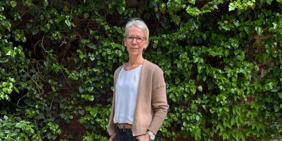 Anja Guth steht im Freien vor einer dichten, grünen Wand aus Efeu. Sie trägt eine beigefarbene Strickjacke über einem weißen Oberteil, eine dunkle, schmal geschnittene Hose und beigefarbene Schuhe. Ihr linker Arm ist locker angewinkelt, die rechte Hand ruht in der Hosentasche. Der Untergrund besteht aus hellem, strukturiertem Boden.