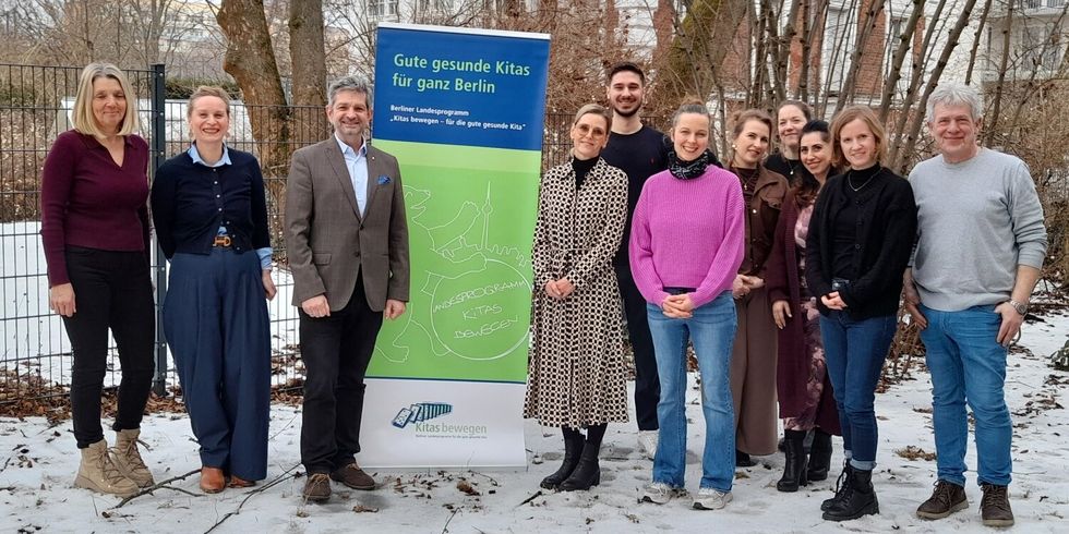 Elf Personen stehen vor einem Banner des Berliner Landesprobramms "Kitas bewegen - für die gute gesunde Kita"