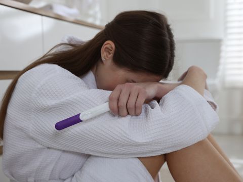 Eine Frau sitzt auf dem Boden, den Kopf auf ihre verschränkten Arme gelegt, wirkt traurig oder besorgt und hält einen Schwangerschaftstest in der Hand.