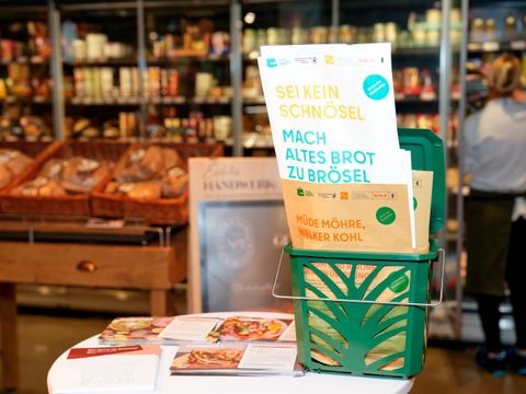 Ein Korb mit Papiertüten auf einem Stehtisch, im Hintergrund ein Supermarktregal