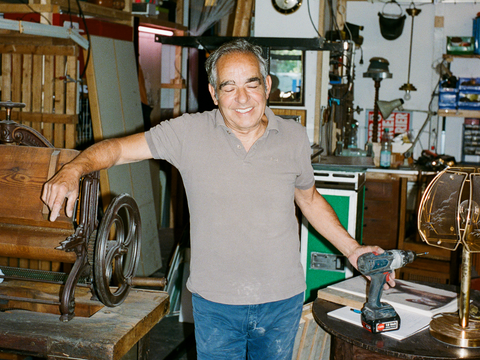 Ein älterer Mann steht in einer Werkstatt. Er hat eine Bohrmaschine in der Hand, im Hintergrund stehen alte Möbel.