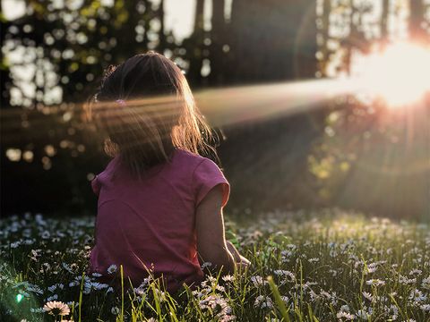 Mädchen von hinten, sitzt auf Blumenwiese, hält ihr Gesicht in einen Sonnenstrahl