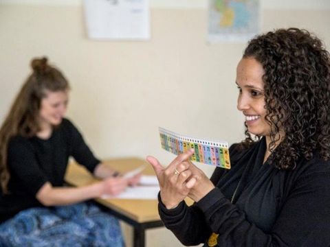 Zwei Frauen im Klassenzimmer; eine zeigt lachend eine farbige Karte, die andere sitzt am Tisch und schaut zu.