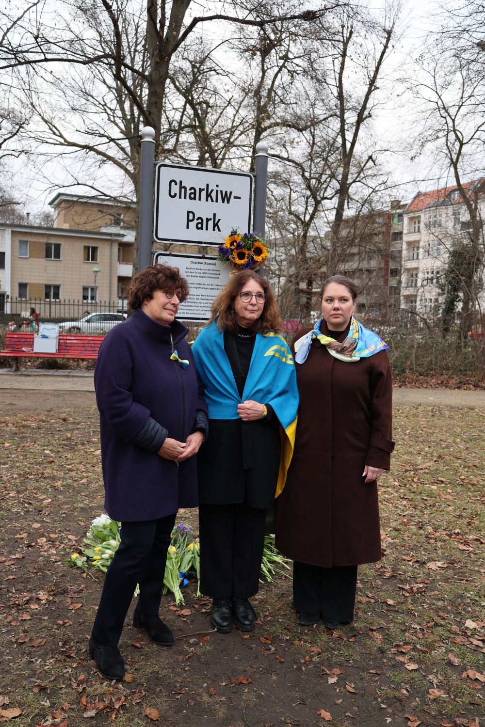 24. Februar 2026: Eingehüllt in Blau-Gelb: Bezirksbürgermeisterin Maren Schellenberg (links) mit Olga Pischel (Vorstandsmitglied des Städtepartnerschaftsvereins; Mitte) und Nina Masol, Botschaftssekretärin der Ukrainischen Botschaft, vor dem Schild des Charkiw-Parks