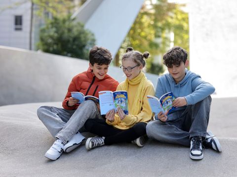 Fotografie von drei lesenden Kindern, die mit Büchern draußen auf dem Boden sitzen