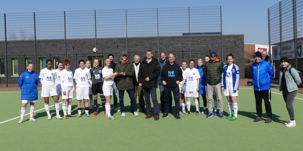 Auf einem Fußballplatz steht eine Gruppe von Menschen zusammen. Manche tragen Sportbekleidung.