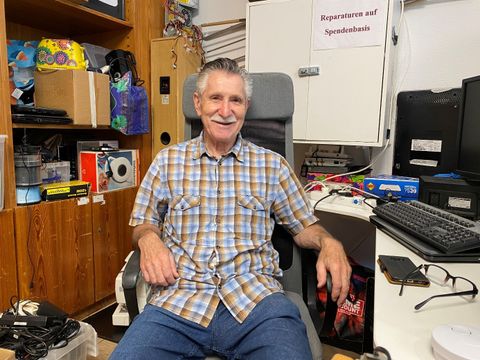 Ein älterer Mann auf Schreibtischstuhl vor Schreibtisch übersäht mit Technik und Ersatzteilen