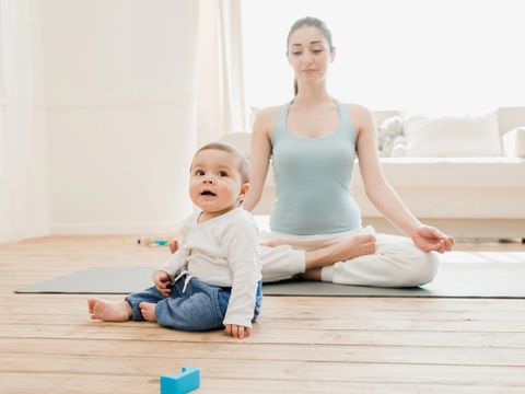 Kleinkind sitzt auf dem Boden, zusammen mit seiner Mutter, die gerade eine Yogapose macht