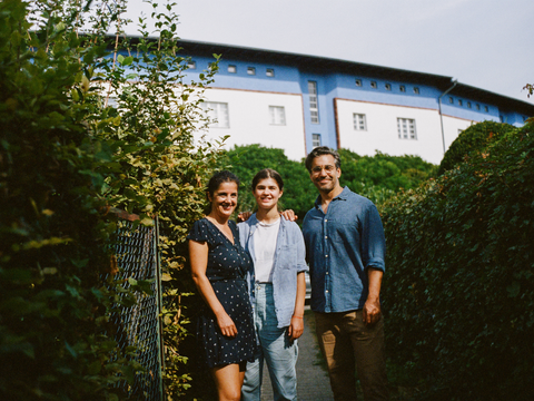 Eine Familie (Mutter, Vater, Tochter) steht zwischen hohen Hecken vor einem Wohnhaus in Neukölln.