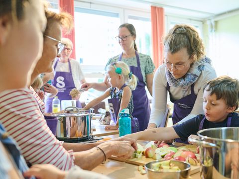 Frauen mit lila Kochschürzen schneiden zusammen mit Kindern Äpfel an einem Tisch
