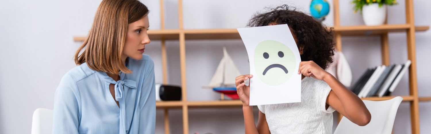 Afroamerikanisches Mädchen mit Autismus bedeckt Gesicht mit unglücklichem Gesichtsausdruck auf Papier in der Nähe von Psychologen auf verschwommenem Hintergrund