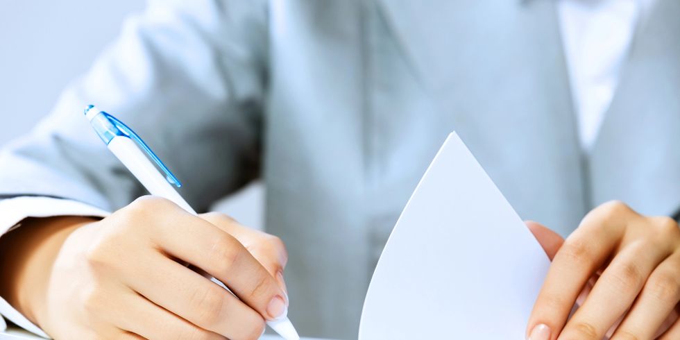 Eine Person in formeller Kleidung hält ein Blatt Papier und schreibt mit einem Stift darauf, vermutlich beim Ausfüllen oder Unterschreiben eines Dokuments.