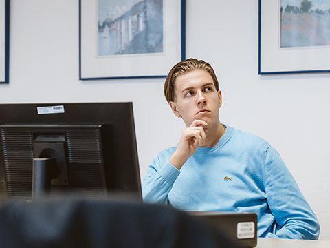 Junger Kursteilnehmer im blauen Pullover sitzt vor PC und schaut interessiert und nachdenklich
