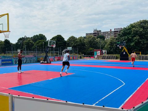 Auf dem Foto sind drei Personen auf einem großen, modernen Outdoor-Basketballplatz mit blauer und roter Spielfläche zu sehen. Im Hintergrund stehen mehrere Basketballkörbe und bunt bemalte Banden mit Städtenamen wie „Dortmund“. Umgeben ist der Platz von grünen Bäumen, einem Zaun mit Graffiti und einem Wohnhaus in der Ferne. Die Atmosphäre wirkt sportlich und sommerlich.
