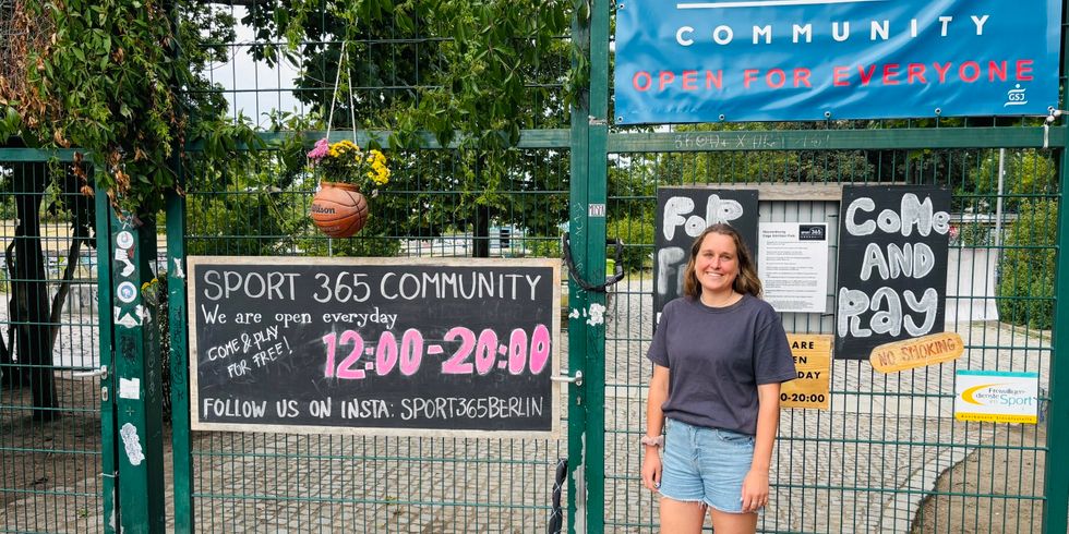 Auf dem Bild sieht man den Eingang eines Sportplatzes mit grünen Gittern und mehreren Schildern, darunter „SPORT 365 COMMUNITY“ und „OPEN FOR EVERYONE“. Die Öffnungszeiten sind von 12:00 bis 20:00 Uhr, und es wird dazu eingeladen, kostenlos mitzuspielen. Eine junge Frau steht rechts vor dem Tor, trägt ein dunkles T-Shirt und Jeansshorts. Das Gelände ist von Bäumen umgeben und wirkt einladend und offen für Besucher.