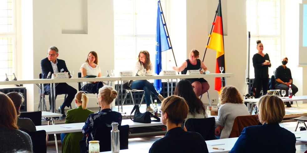 Vier Menschen sitzen nebeneinander an Tischen auf einem Podium. Daneben steht eine Gebärdendolmetscherin. Mehrere Personen sitzen zum Podium gewendet an weiteren Tischen.