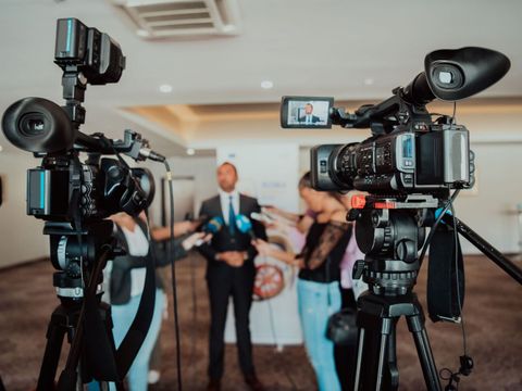 Zwei Kameras stehen in einem Raum und sind auf einen Mann gerichtet, der von mehreren Reportern interviewt wird.