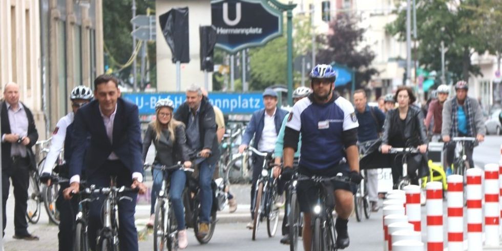 Das Bild zeigt mehrere Fahrräder auf der neu eröffneten gesicherten Fahrradspur entlang der Karl-Marx-Straße. Am rechten Rand stehen weiß-rot gestreiften Poller, die die Fahrradspur von der Straße abtrennen. Im Hintergrund sind blaue Schilder des Eingangs zum U-Bhf. Hermannstraße zu sehen. An der Spitze der Fahrradgruppe fährt Bezirksbürgermeister Martin Hikel.