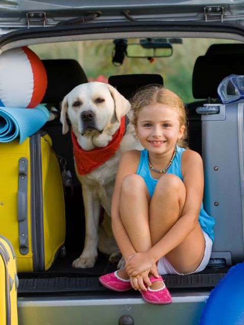 Ein kleines Mädchen und ein Hund sitzen neben Koffern im offenen Kofferaum eines PKW´s