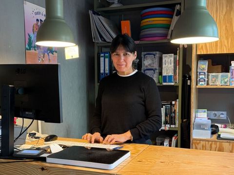 Mohidil Shamshidinova steht hinter einem hölzernen Tresen in der Else-Ury-Bibliothek. Auf dem Tresen befinden sich ein Computerbildschirm, eine Tastatur und ein Tablet. Zwei große, grüne Pendelleuchten hängen von der Decke und beleuchten den Arbeitsbereich. Im Hintergrund sind Regale mit Büchern, Ordnern und bunten Gegenständen zu sehen. Rechts neben dem Tresen stehen mehrere Kisten und ein Bücherwagen. Die Wände und Möbel bestehen überwiegend aus hellem Holz.