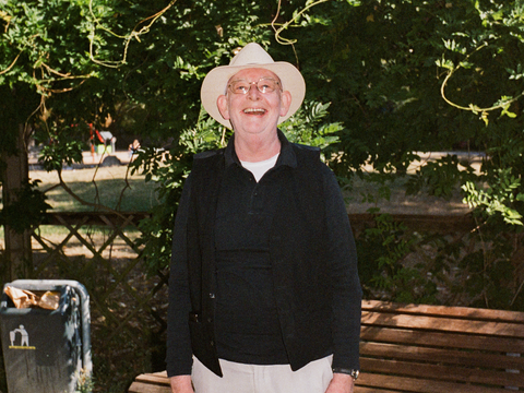 Ein älterer Herr mit Sonnenhut steht vor einer Parkbank und lacht in die Kamera.