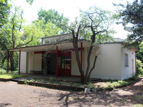 Versuchspavillion von Bruno Taut und Fritz Karsen: weißer Bungalow mit erhobenem Dach mit Fensterfront. Großer Eingangsbereich mit überdachter Terrasse