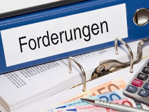 Auf einem geöffneten Aktenordner liegen ein Taschenrechner, ein Stift, mehrere Geldscheine und weitere Ordner mit der Aufschrift 'Forderungen'