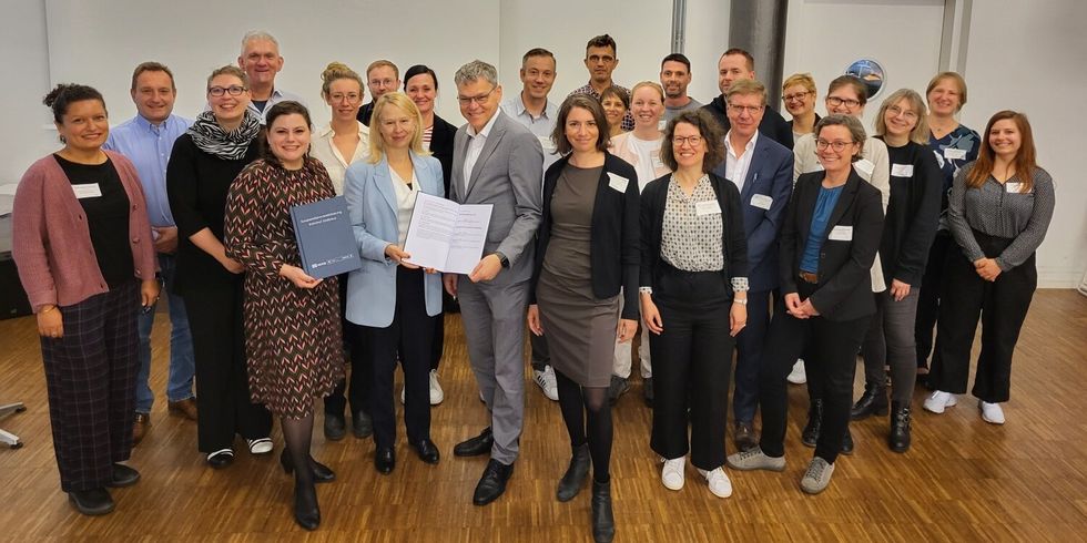 Eine Gruppe Menschen steht zusammen in einem Saal. Die Personen im Vordergrund halten eine Kooperationsvereinbarung in den Händen.