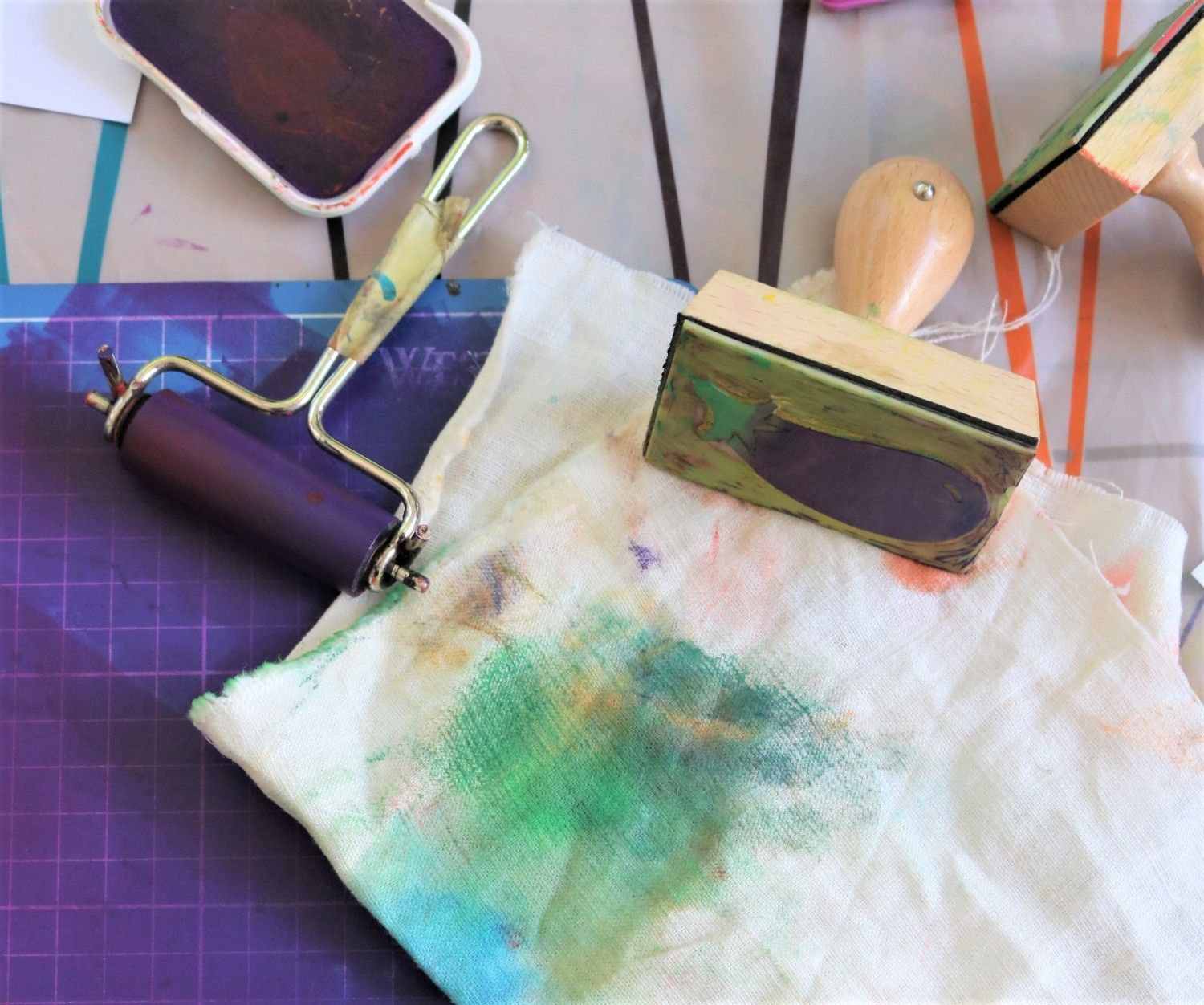 Ein weißes Tuch mit grünen und blauen Tuschflecken. Auf dem Tuch liegt ein großer Holzstempel mit einem Wal als Stempelmotive. Links neben dem Tuch liegt eine blaue Farbrolle.