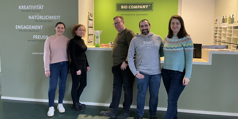 Fünf Personen stehen nebeneinander an einer Theke. Dort steht ein kleiner weiß-grüner Buddy-Bär. Im Hintergrund an der Wand ist das Logo der Bio Company.
