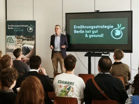 Ein Mann hält eine Rede, man sieht mehrere Zuschauerreihen von hinten, neben dem Redner steht ein Smartboard, auf welchem das Logo der Berliner Ernährungsstrategie zu sehen ist