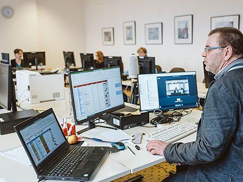 Kursszene EDV, Kursleiter sitzend am Tisch mit drei Monitoren; im Hintergrund Kursteilnehmende.