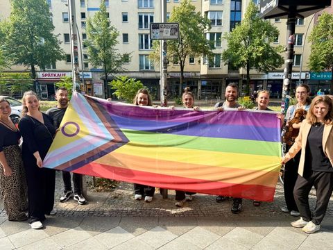 „Gemeinsam anders“ lautet das Motto des Bezirksamtes Friedrichshain-Kreuzberg und so darf auch die „Progress-Flag“ anders herum präsentiert werden: Bezirksbürgermeisterin Clara Hermann (re.) und Mitarbeiter*innen vor dem Rathaus Friedrichshain-Kreuzberg.