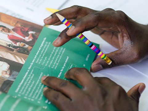 Dunkelhäutige Hand hält einen Stift, davor liegt ein geöffnetes Buch