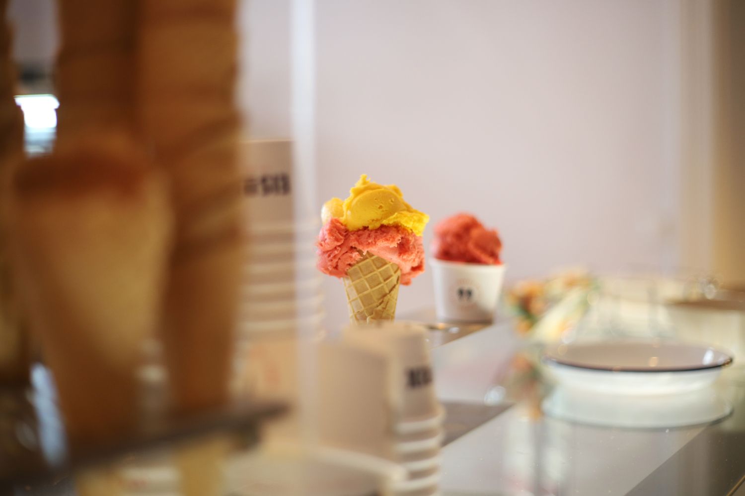 Im Vordergrund auf der linken Bildseite mehrere gestapelte Eiswaffeln. In der Mitte des Bildes eine Eiswaffel mit zwei Kugel Eis drin. Die untere Kugel ist hell rosa, die obere Kugel ist gelb. Dahinter noch ein weißer Papp-Eisbecher mit einer dunkelroten Kugel Eis darin. Alle steht auf einer gläsernden Theke.