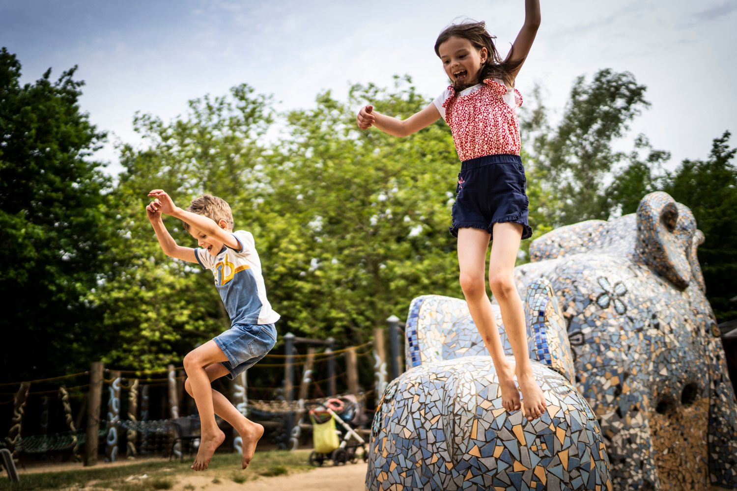 Ein Mädchen und ein Junge springen von einem mit Mosaiken verzierten Spielgerät. Das Mädchen trägt ein rot-rosa-weißes Top und blaue kurze Hosen. Der Junge trägt eine knielange Jeans und ein T-Shirt mit einem weißen, gelben und blauen Streifen.