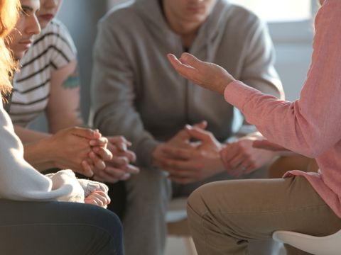 Eine Gruppe von Personen sitzt in einem Kreis und hält die Hände gefaltet. Eine Person spricht und gestikuliert mit einer Hand, während die anderen aufmerksam zuhören, typische Szene einer Gruppentherapie oder Besprechung.