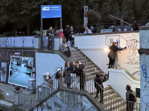 Man sieht eine Treppe, die zum Wasser nach unten führt Auf der Treppe sind mehrere Personen zu sehen. Eine Person FRau steht auf dem Zwischenpodest der Treppe auf einer Leiter, die an die Wand gelehnt ist. Sie malt mit einem schwarzen Stift etwas auf die weiße Wand.