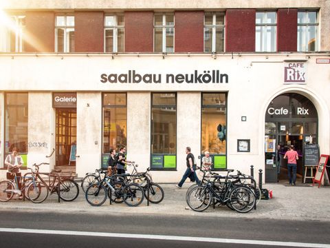 Außenansicht der Galerie Saalbau von der Straßenseite aus. Es ist eine helle Hausfassade in Erdgeschosshöhe zu sehen. Einzelne hohe Fenster unterbrechen die Fassade. Oberhalb der Fenster ist der aus schwarzen einzelnen Buchstaben bestehende Schriftzug "saalbau neukölln" zu lesen. Auf dem Bürgersteig vor dem Haus stehen viele Fahrräder angelehnt an Fahrradbügel. Es laufen mehrer Personen zwischen Fahrrädern und Hausfront entlang. Die Sonne scheint.