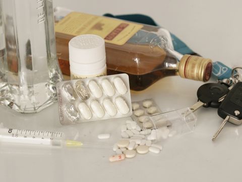 auf einem weißen Tisch liegende Weinbrandflasche mit umliegenden Tabletten (mit umgekippten durchsichtigen Döschen) und einer weiteren Dose, einer Wodkaflasche sowie einen Autoschlüssel am Schlüsselband
