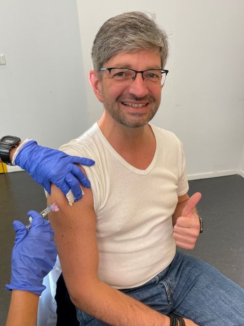 Ein Mann sitzt auf einem Stuhl und macht den Daumen hoch. Er wird von jemanden mit OP-Handschuhen in den Arm geimpft.