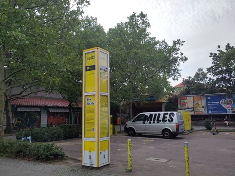 eine gelbe Informationssäule, ein Lieferwagen des Carsharingdiensts "MILES", eine Ladestation für elektrische Carsharingfahrzeuge und 2 leihbare E-Tretroller.