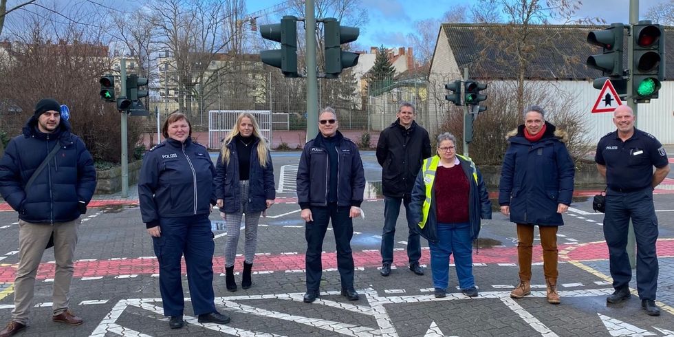 Acht Menschen stehen auf einer kleinen Straße mit Ampeln und Schildern. Manche tragen blaue Jacken mit dem Schriftzug "Polizei".
