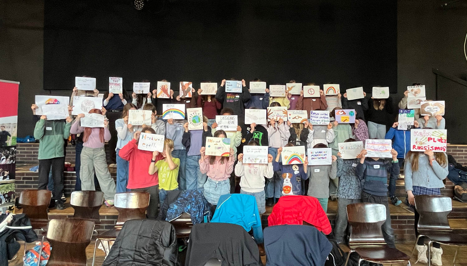 Viele junge Menschen stehen auf der Bühne und verdecken ihr Gesicht mit davorgehaltenen Plakaten
