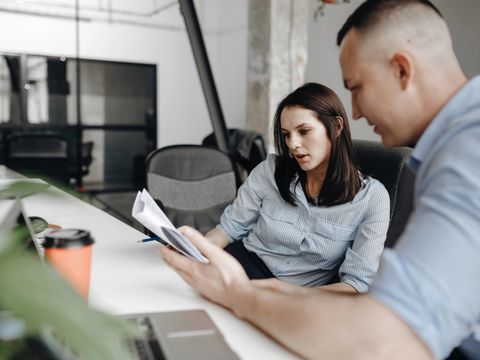 Zwei Personen, Frau und Mann, in Bürokleidung arbeiten gemeinsam am Schreibtisch sitzend mit Dokumenten in einem hellen, modernen Büro