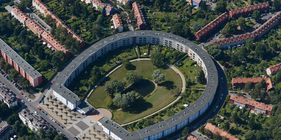 Luftbildaufnahme der Hufeisensiedlung. In der Mitte des Bildes die Hufeisensiedlung mit einem grauen Dach. Der Innenhof der Siedlung ist begrünt und es gibt einen kleinen Teich in der Mitte. Rechts und links, sowie oberhalb der Hufeisensiedlung gehen strahlenförmig weitere Reihen Häuser weg, von denen nur die roten Dächer zu sehen sind
