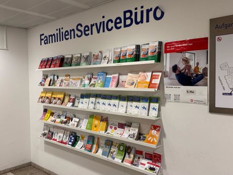 Vor dem Eingang des FamilienServiceBüro Friedrichshain-Kreuzberg befindet sich eine Wand mit mehreren Reihen von Prospekthaltern. In den Haltern stecken zahlreiche Broschüren und Flyer in unterschiedlichen Farben und Formaten. Über den Prospekten ist der Schriftzug 'FamilienServiceBüro' in großen, blauen Buchstaben an der Wand angebracht.