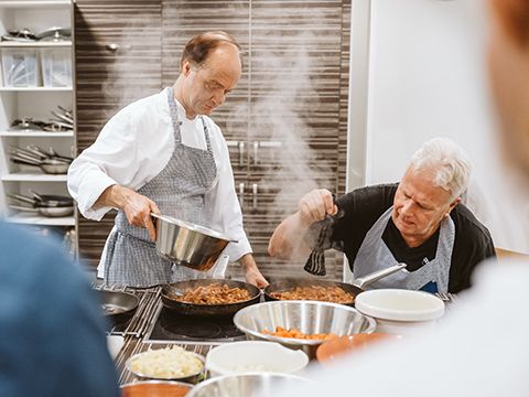 Kochkurs mit Kay Ivers - Kochszene mit Kursleiter und einem sitzenden Mann, der Gewürze in die Pfanne streut