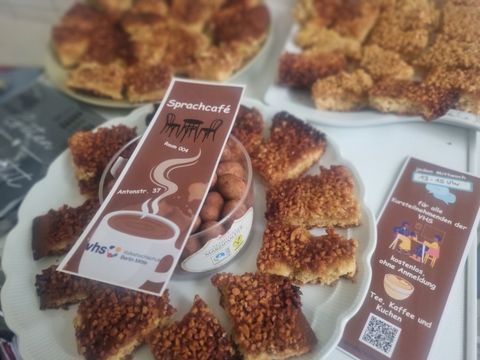 Teller mit Kuchen und Infozetteln für ein Sprach-Café. Auf einem Zettel steht: Sprachcafé, Raum 004, Antonstraße 37. Dazu gibt es Tee, Kaffee und Kuchen.